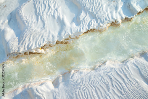 Hot mineral water stream running over textured white travertine surface in Pamukkale, Turkey. Close-up view showing natural erosion patterns and subtle color variations under soft daylight.
