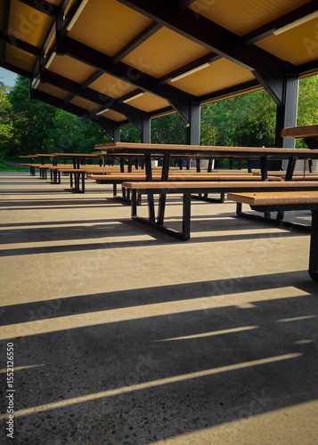 a covered outdoor pavilion with multiple picnic tables and benches, illuminated by natural light casting long shadows across the concrete ground. park architecture