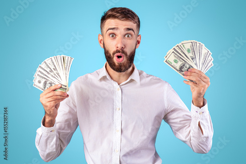 Shocked young man holding money banknotes on blue studio background. Caucasian guy cannot believe his luck, excited over big win or financial success, happy over raise of income or salary