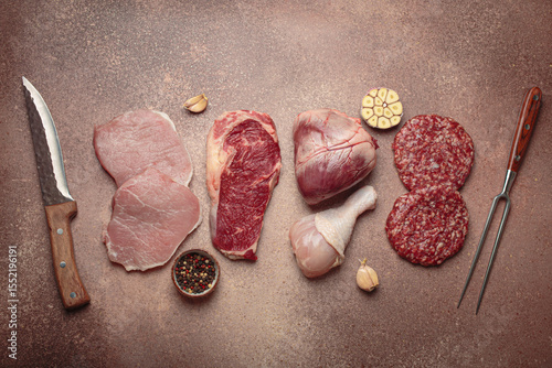 Selection of assorted raw meat cuts for zero carb carnivore diet: uncooked beef steak, pork, ground meat patty, heart, and chicken legs on beige stone background top view with knife and carving fork