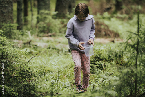 Fotografie A young girl in a lavender hoodie and pink pants walks through a mossy forest, holding a plastic jar, possibly collecting insects or exploring nature