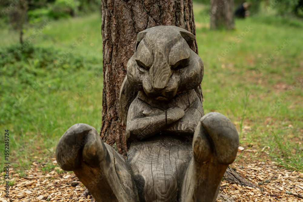 Obraz premium funny wooden rabbit sculpture in forest park, carved bunny sitting with arms crossed on woodchips, whimsical outdoor woodland artwork for kids area, humorous handmade animal statue in nature