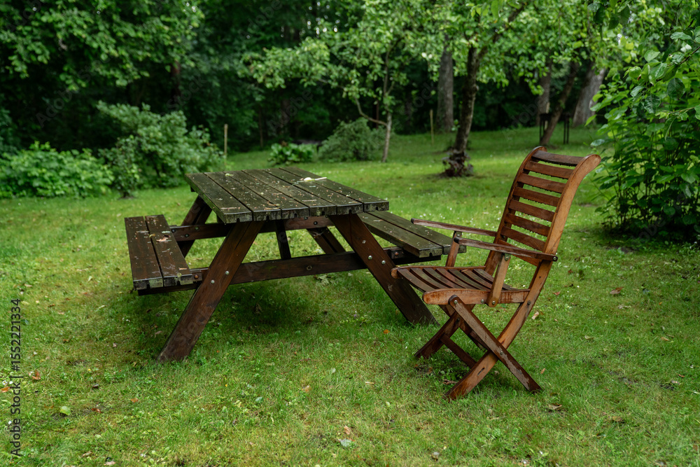wooden picnic table with bench and garden chair on green grass in backyard with trees, outdoor rustic furniture for relaxing and dining in summer garden, countryside setting with natural greenery wooden picnic table with bench and garden chair on green grass in backyard with trees, outdoor rustic furniture for relaxing and dining in summer garden, countryside setting with natural greenery