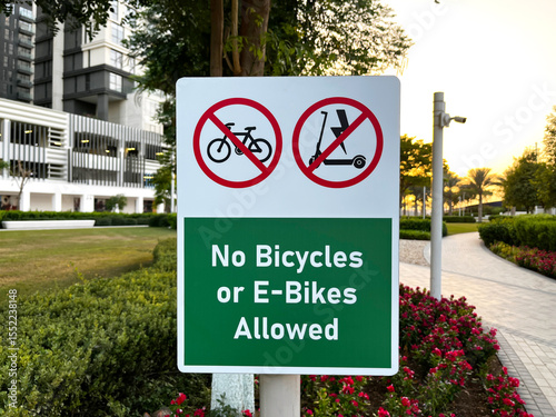 Urban park sign prohibiting bicycles and e-bikes, with flowers, walkway, and modern buildings at sunset.