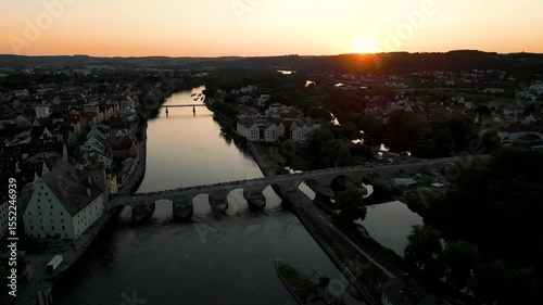 Regensburg at sunset captured from above, featuring the iconic Stone Bridge over the Danube River and the majestic twin towers of St. Peter's Cathedral glowing in warm evening light