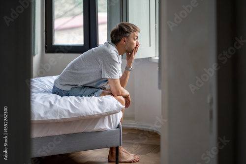 Tired man sits on bed lost in deep thought after sleepless night. Exhausted male suffering from insomnia, feeling lonely, overthinking problems, dealing with relationship breakup, midlife challenges.