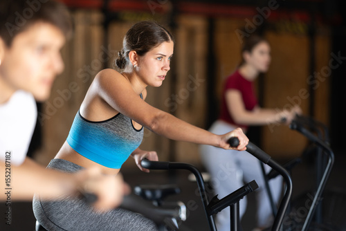 In gym, girl does exercises using airbike. Young group of athletes trains in gym. Physical education and sports, active healthy lifestyle.