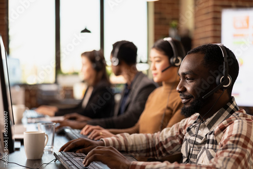 Closeup of african american call center agent reviewing information on his desktop pc while consulting with remote customers. Modern workspace with multiethnic telemarketers using their computers.