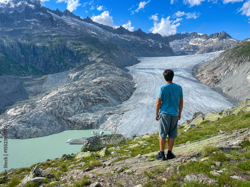 Naklejka premium Man stands admiring the stunning view of vast river of ice flowing into a turquoise glacial lake. Dramatic mountain peaks rise majestically above Rhone Glacier, accentuating the grandeur of landscape.