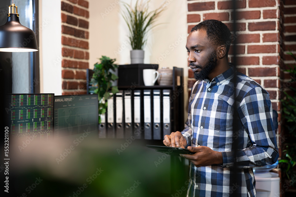 Fototapeta premium African american man with tablet, working night shift, examining financial reports on computer screen in modern workspace. Black male manager holding digital device as he reviews stock on desktop pc.