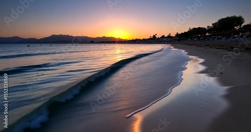 Coastal sunset video showing golden sun setting over a calm sea with gentle waves and a sandy beach under a twilight sky