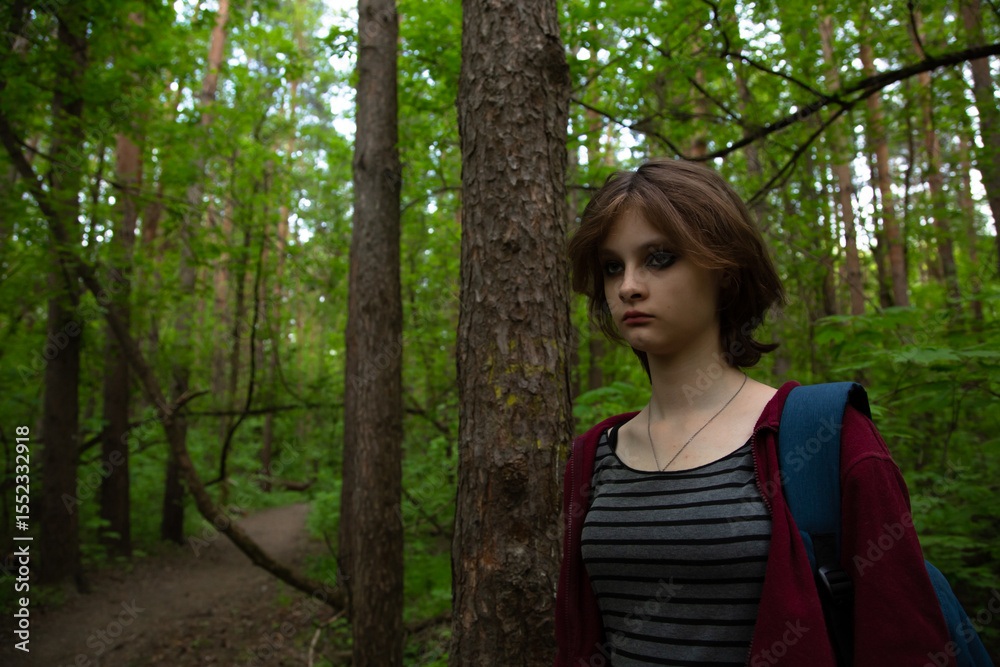 Beauty face young woman with serious thinking look hiking among green pine trees on the path in the forest with blue backpack on shoulder. Girl walking in woods, in fashion clothes Emotion portrait Beauty face young woman with serious thinking look hiking among green pine trees on the path in the forest with blue backpack on shoulder. Girl walking in woods, in fashion clothes Emotion portrait