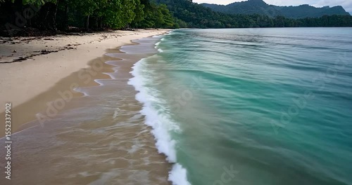 Video of a tropical beach with turquoise water white sand and lush green vegetation along the shoreline waves gently rolling in on a sunny day