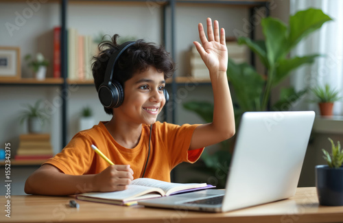 Young Indian kid in online class at home. Boy using laptop, smiles, raises hand, and studies. E-learning concept, remote education, digital learning. Happy child in headset studies remotely.
