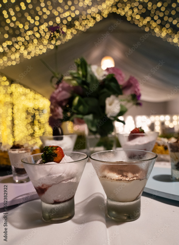 Naklejka premium Catering service for a birthday. Closeup view of the desserts table with modern tiramisu and cheesecake glass cups with a beautiful floral arrangement. The garden lights and tent in the background