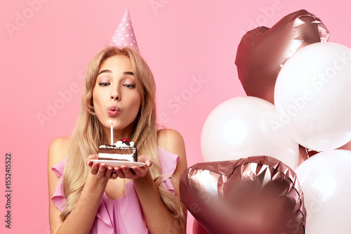 Canvas Print Cute young lady with birthday hat and balloons blowing candle on piece of cake over pink studio background, panorama with copy space
