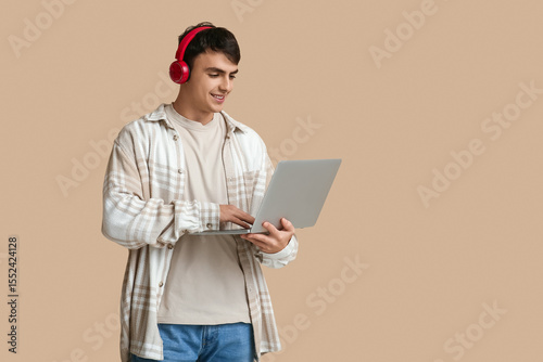 Handsome young man with headphones and laptop on beige background