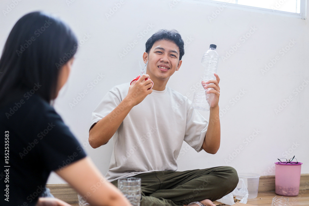 Fototapeta premium Smiling Young Asian Male Showing Scissors And Plastic Bottle While Sitting On Floor
