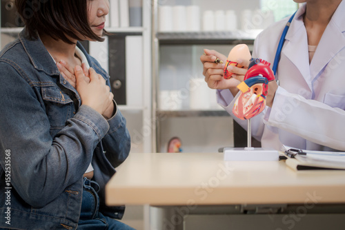 A young Asian female doctor consults with a patient seated across the desk. Using a heart model, she explains cardiovascular health conditions, offering guidance and support in calm clinical setting