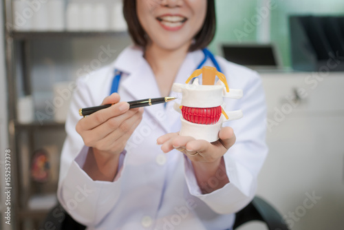 A middle-aged Asian female doctor sits at her desk, pointing to a spinal model with a highlighted red section. She explains spinal conditions like herniated discs or nerve compression to a patient