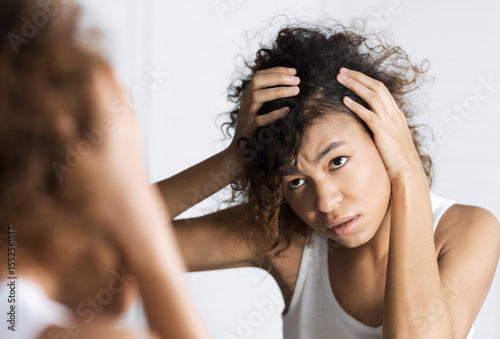 Afro-american girl frowning and looking intently on her face in mirror. Hair and sculp problems concept