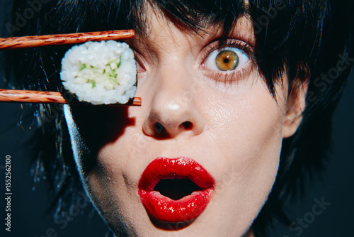 Young woman expressing surprise while holding sushi with chopsticks, vibrant makeup, dark background.