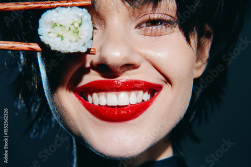 Young woman with dark hair holding sushi, showcasing a bright smile and red lipstick, perfect for food and lifestyle imagery.
