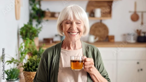 A smiling elderly woman with white hair enjoys a cup of tea in a cozy kitchen with plants in the background.