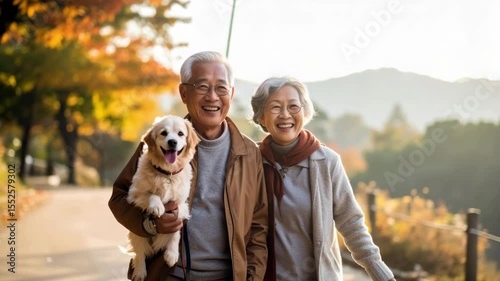 An elderly couple walks outdoors on a sunny day, smiling and holding a fluffy dog, with autumn trees in the background.
