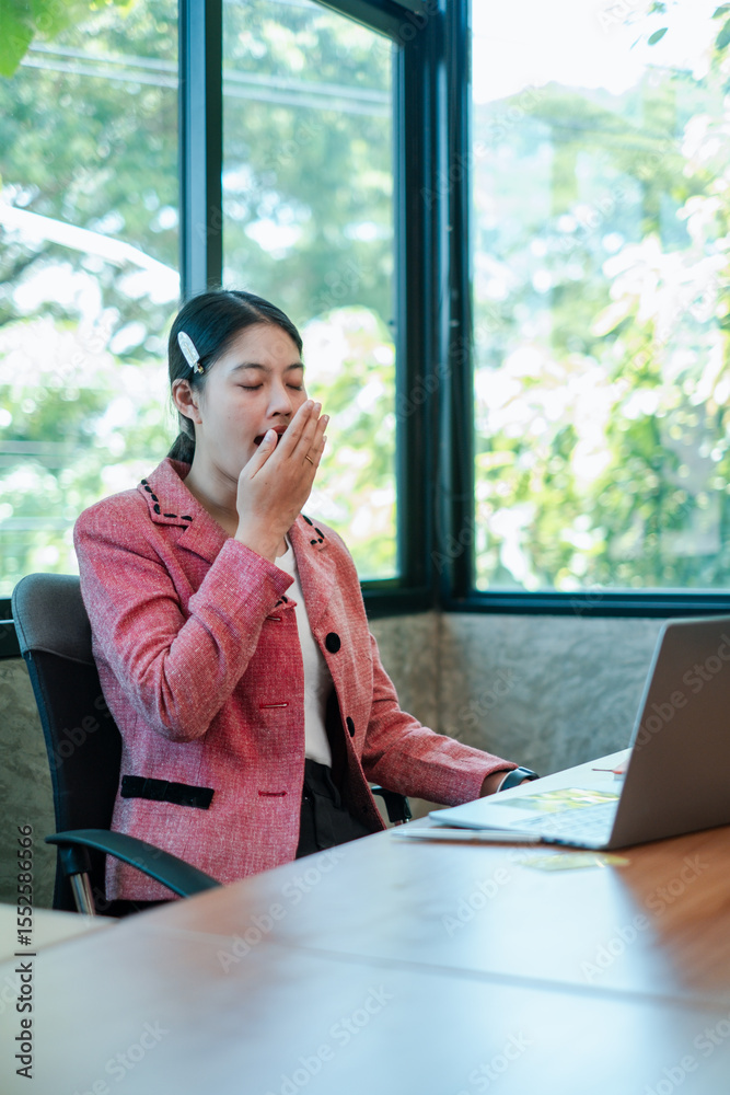 A professional woman in a pink blazer yawns at her desk in a bright office with a laptop and large windows. A professional woman in a pink blazer yawns at her desk in a bright office with a laptop and large windows.