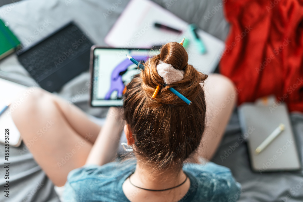 Naklejka premium Top view of girl sitting cross-legged on bed with stylus and tablet, painting colorful cartoon illustration. Creative and casual atmosphere with work tools around.