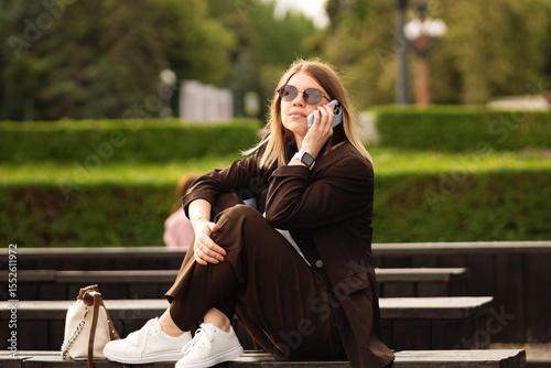 Young woman in brown suit sitting and using smartphone outdoors. Great natural vibe and showcase stylish, relaxed mood. Outfit looks effortlessly chic