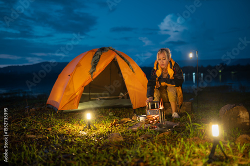 A woman is sitting by a campfire. She is wearing a yellow jacket. There is a tent nearby