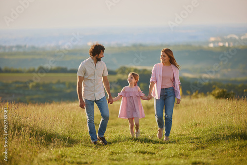 Obraz na plátně Family takes a leisurely stroll in a green field, holding hands and sharing smiles, while enjoying a warm afternoon under the sun