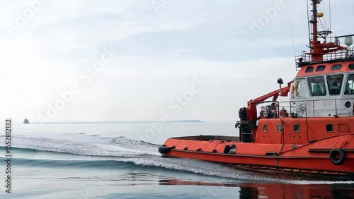 Vibrant red tugboat navigates through calm waters, gently churning the surface as waves ripple outward, maritime operations unfolding in the distance under a clear white sky, Dynamic Camera