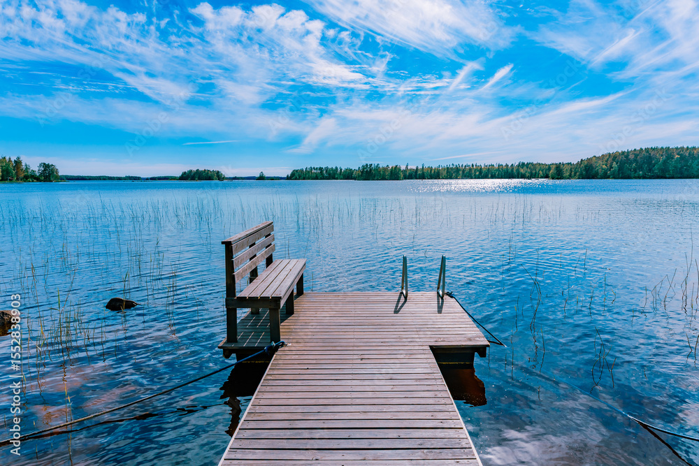 Obraz premium Wooden pier on blue lake with a bench and a ladder in Finland. The water is calm and the sky is blue