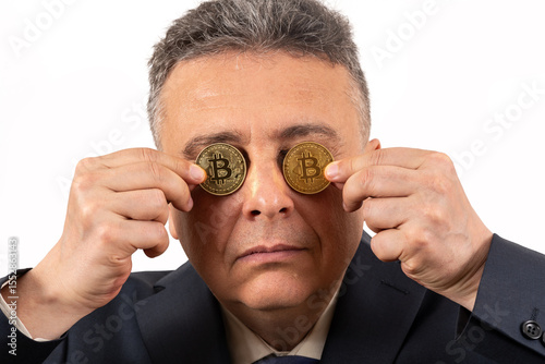 A middle-aged man in a business suit hides his eyes behind coins with the Bitcoin cryptocurrency symbol