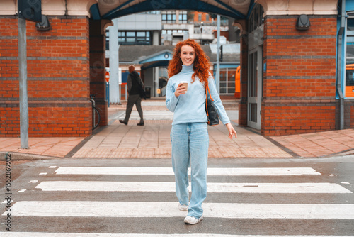 Young woman with curly red hair walks confidently across a city street, enjoying coffee in casual outfit during a sunny day in an urban area