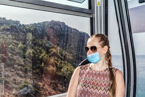Woman wearing a face mask and sunglasses enjoying a scenic ride on the Alanya Teleferik cable car. The background reveals a mountainous landscape and distant sea views
