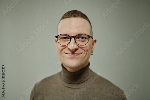 Portrait of young adult Caucasian man with short hair, wearing glasses and nose ring, smiling while looking directly at camera against plain background