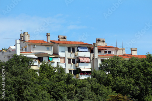 Cluster of white apartment buildings with red tile roofs and balconies rises above dense green treetops, featuring colorful awnings and multiple chimneys under a clear blue sky