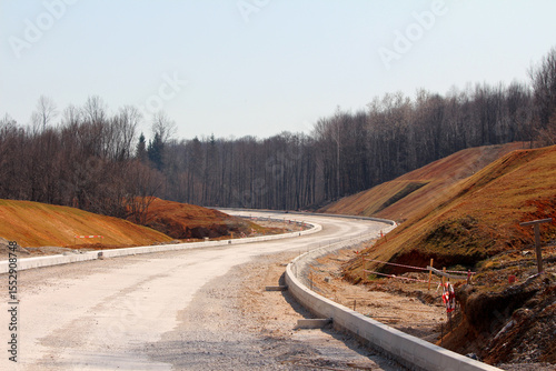Wide gravel road under construction curves through a hilly landscape with orange-brown slopes, bordered by concrete curbs and sparse safety markers, with leafless forest in the background