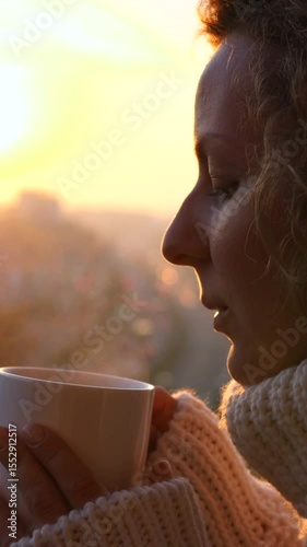 Vertical screen golden hour sunlight illuminates a woman wearing a knitted sweater, savoring a hot drink and appreciating the urban landscape