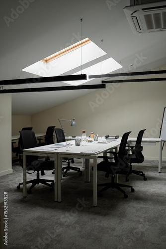 Empty modern office workspace featuring several black office chairs surrounding desks under skylight, various office supplies and coffee cups scattered on tables, no people present