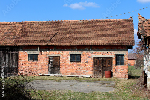 Rectangular outbuilding made of exposed red bricks features a steep tiled roof, two small windows, and weathered wooden doors, set in a grassy yard under a clear blue sky