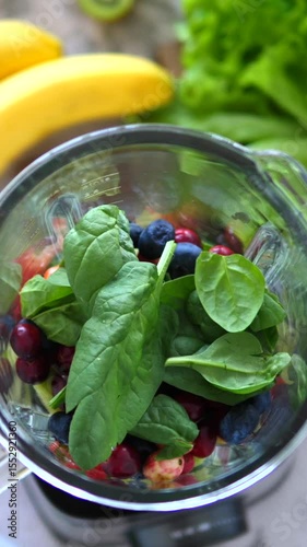 Vertical screen fresh spinach leaves are added to a blender already containing mixed berries and chopped fruits, ready to be blended into a healthy and refreshing smoothie