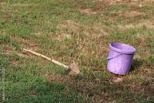 Worn purple plastic bucket and a metal-bladed hoe with wooden handle rest on uneven grassy ground, suggesting recent gardening or fieldwork activity under bright sunlight