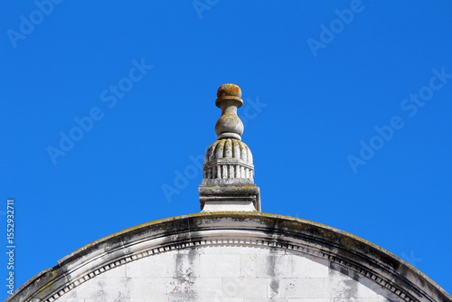 Ornate stone finial with weathered details sits atop a curved building cornice, highlighted against a clear blue sky, showing classical architectural style and aged surface