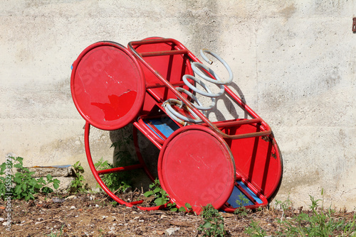 Red metal playground spring rider with circular seats and white coil spring lies overturned on soil and weeds, leaning against a weathered concrete wall in sunlight