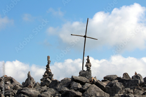 Simple wooden cross stands among balanced rock cairns on a rocky hilltop, set against a bright blue sky with scattered clouds, symbolizing faith and marking a summit or memorial site